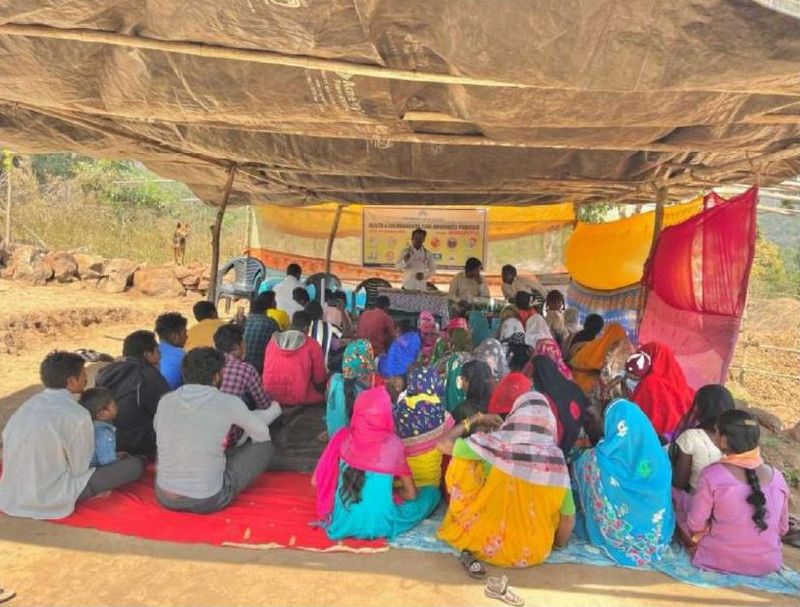 A community awareness session held under a tarpaulin shelter, with villagers seated on the ground